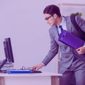 Image shows a man, in an office, wearing a suit and holding a yoga mat. He is logging off the computer to go work out. Image has a purple filter on it | fit4mii